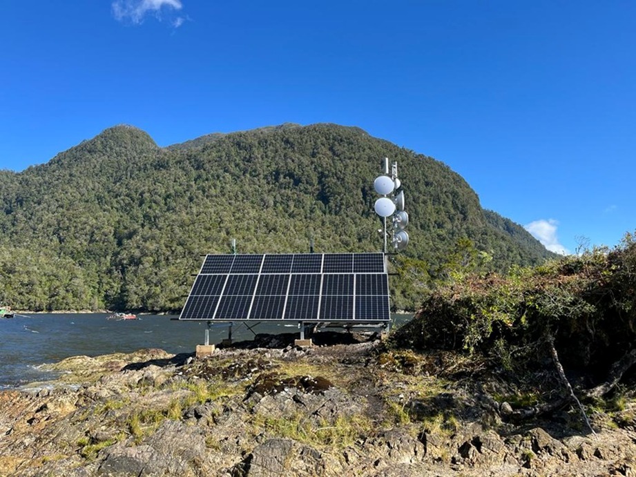 Radio Lodge™ instalado en costa patagónica con paneles solares y torre de telecomunicaciones, rodeado de montañas boscosas